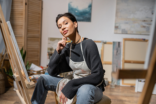 Brunette artist holding paintbrush and looking at camera near blurred canvases in workshop