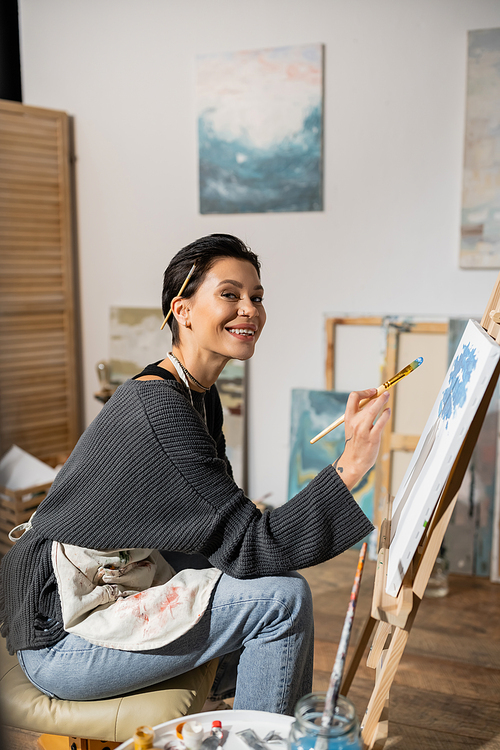 Positive artist looking at camera while holding paintbrush near canvas in studio