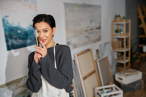 Smiling artist in apron holding paintbrushes near face in studio