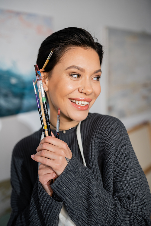 Portrait of positive artist holding paintbrushes and looking away in studio