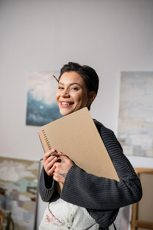 Portrait of cheerful artist in apron looking at camera while holding sketchbook