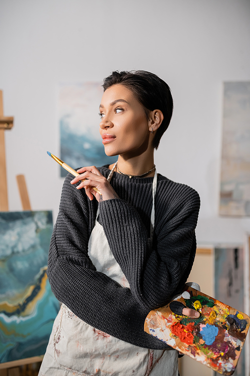 Short haired artist in sweater and apron holding palette with paints and paintbrush