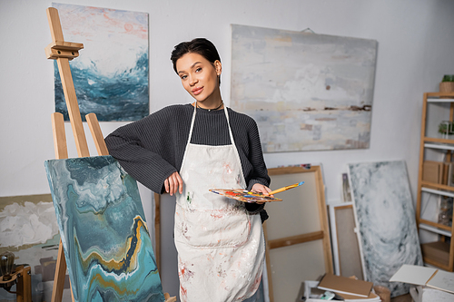 Brunette artist holding paintbrush and palette near canvas on easel in studio