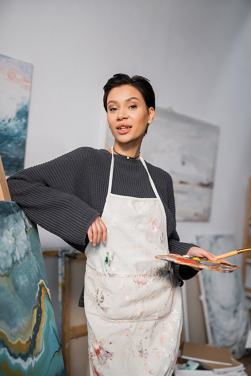 Brunette artist in dirty apron holding palette near drawing in workshop
