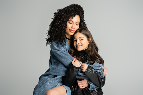 Curly woman in denim jacket hugging happy daughter isolated on grey