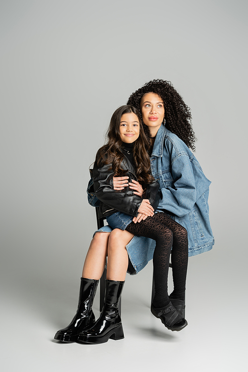 Stylish woman holding smiling daughter while sitting on grey background