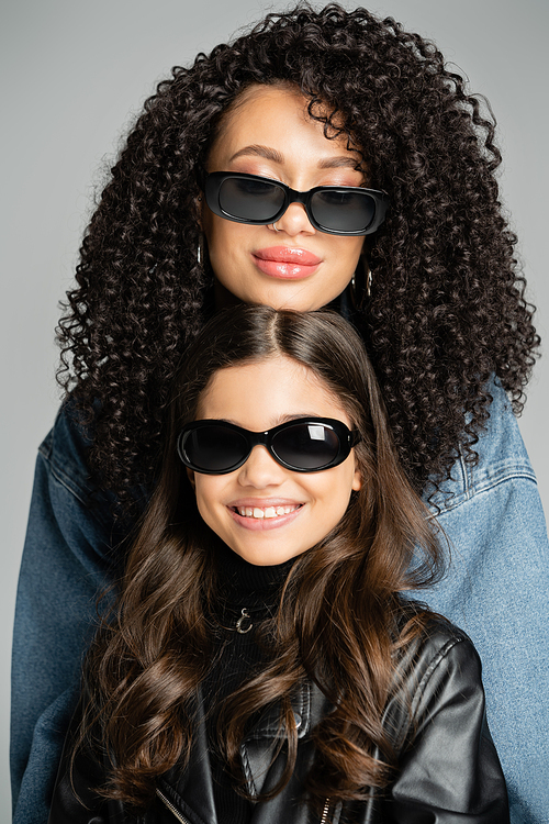 Portrait of curly woman in sunglasses posing near smiling daughter isolated on grey