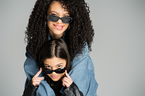 Cheerful woman in sunglasses and denim jacket hugging daughter isolated on grey