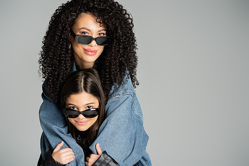 Smiling woman in sunglasses hugging preteen daughter isolated on grey