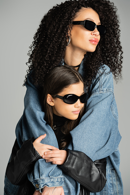 Fashionable woman in sunglasses hugging daughter isolated on grey