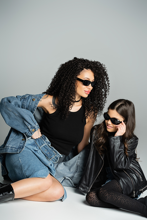 Stylish woman in denim jacket and sunglasses sitting near daughter on grey background