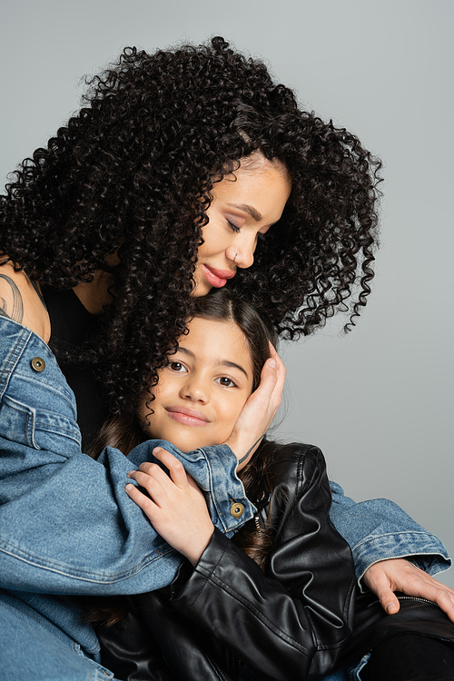 Curly mother hugging smiling daughter in leather jacket isolated on grey