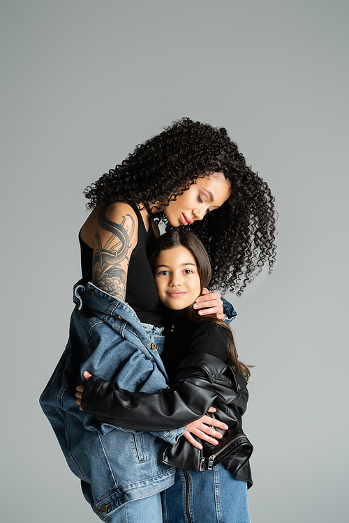 Tattooed mother in denim jacket hugging daughter isolated on grey