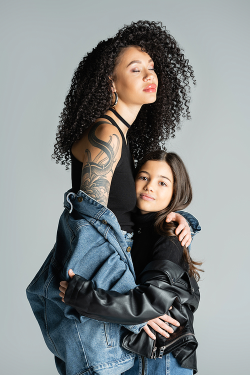 tattooed young woman in denim jacket posing with child isolated on grey