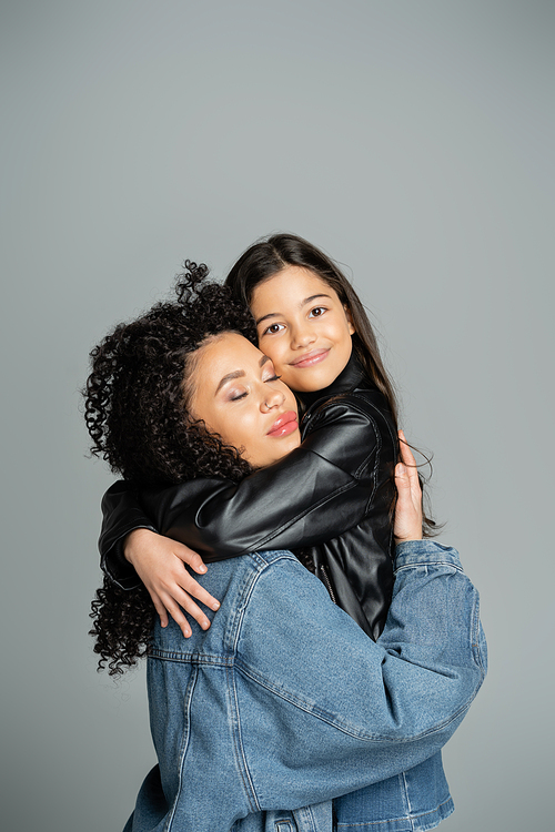 Curly woman in denim jacket hugging cheerful preteen daughter isolated on grey