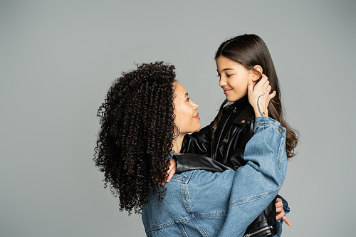 Stylish curly woman in denim jacket touching happy daughter isolated on grey