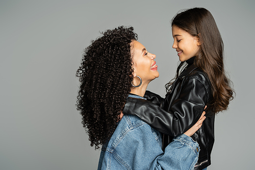 Side view of cheerful mom in denim jacket sticking out tongue while hugging daughter isolated on grey