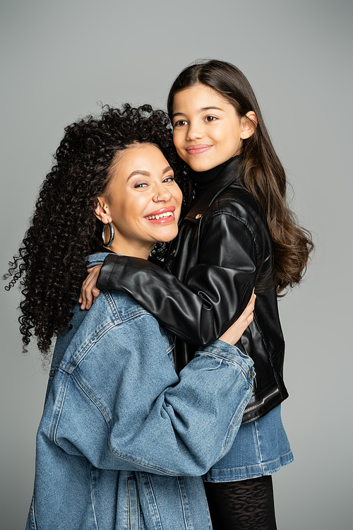 Smiling curly mother in denim jacket hugging stylish child isolated on grey