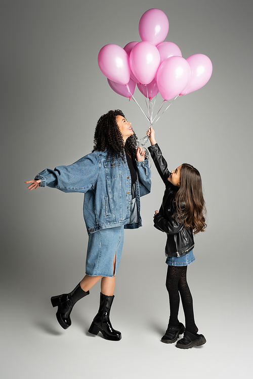 Side view of stylish mother and daughter holding pink balloons on grey background