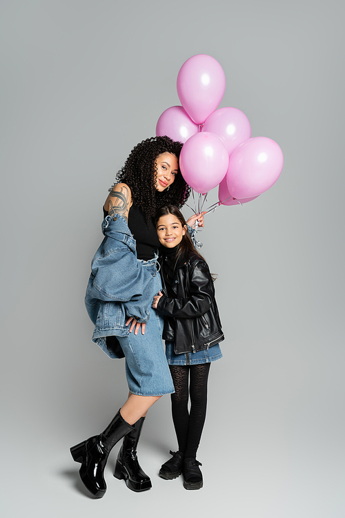 Full length of tattooed woman holding pink balloons near daughter on grey background