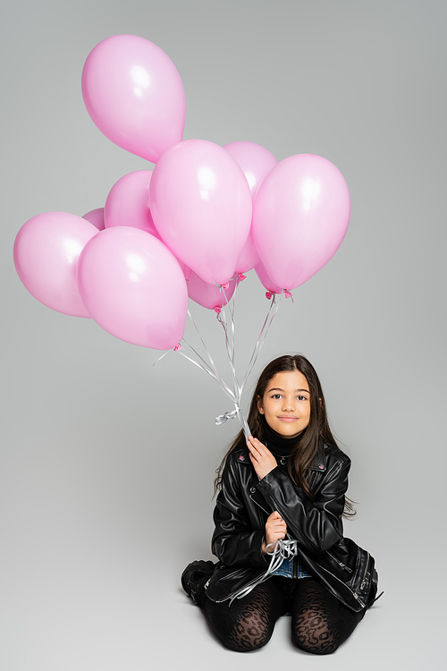 Stylish kid in leather jacket holding pink balloons on grey background