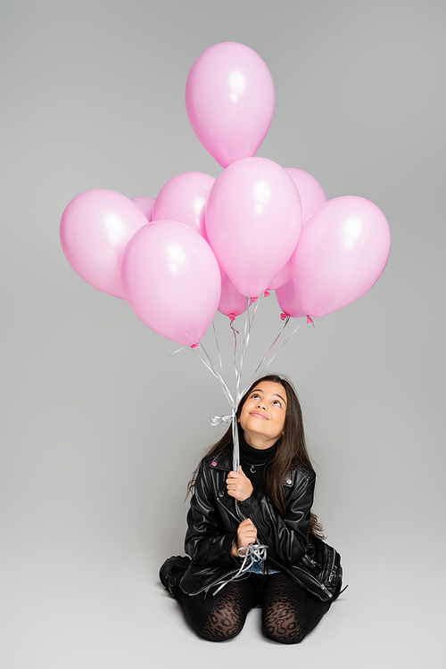 Cheerful and stylish girl looking at pink balloons on grey background