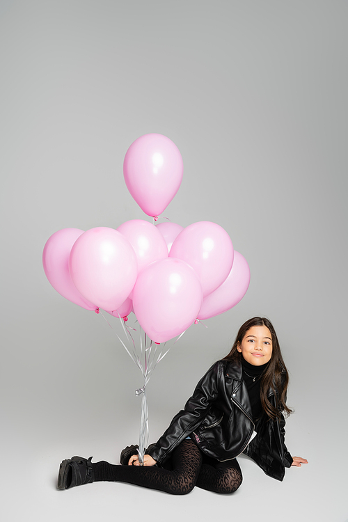 Smiling preteen kid in leather jacket looking at camera near pink balloons on grey background
