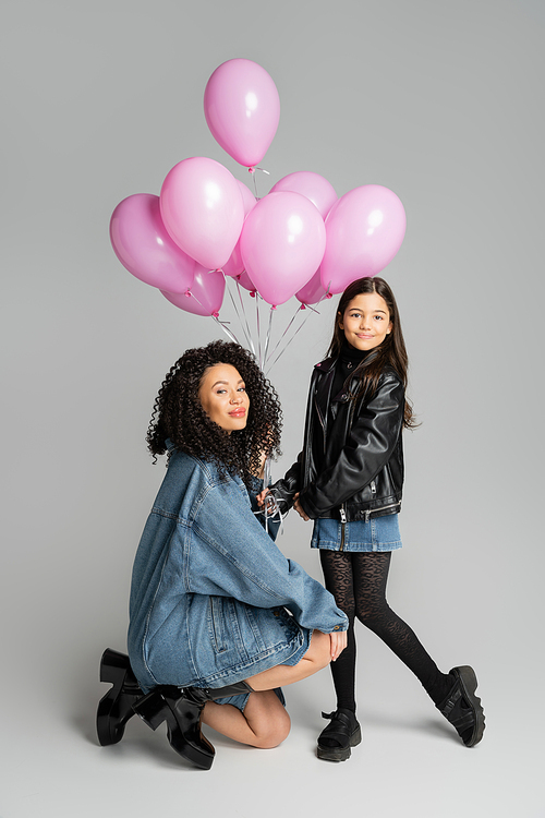Fashionable woman looking at camera near daughter and pink balloons on grey background