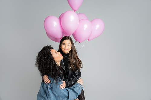 Curly woman hugging smiling daughter near pink balloons isolated on grey