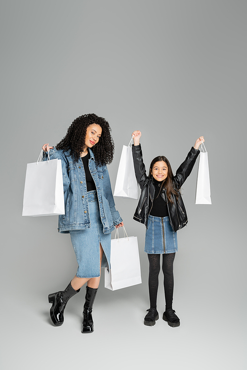 Full length of stylish mother and daughter holding shopping bags on grey background