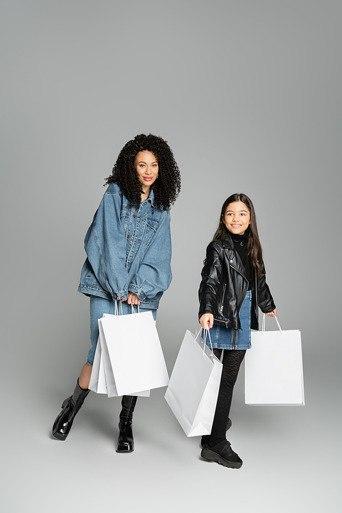 Full length of trendy young woman holding shopping bags near daughter on grey background