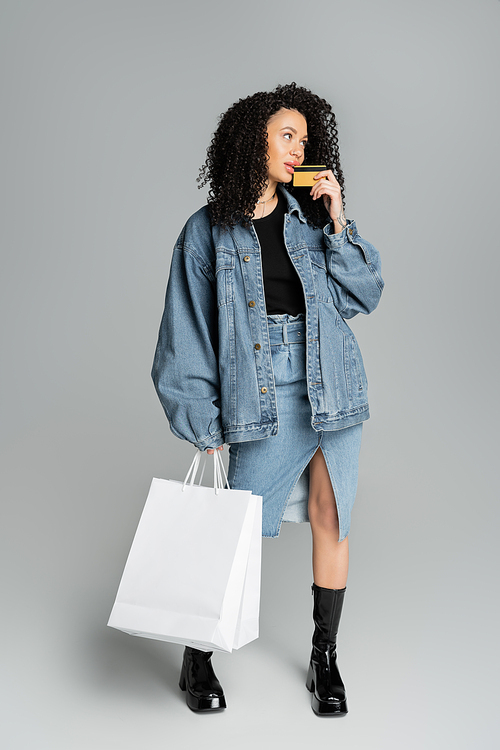 Full length of stylish woman holding credit card and shopping bags on grey background