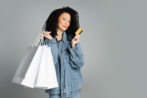Trendy woman in denim jacket holding credit card and shopping bags isolated on grey