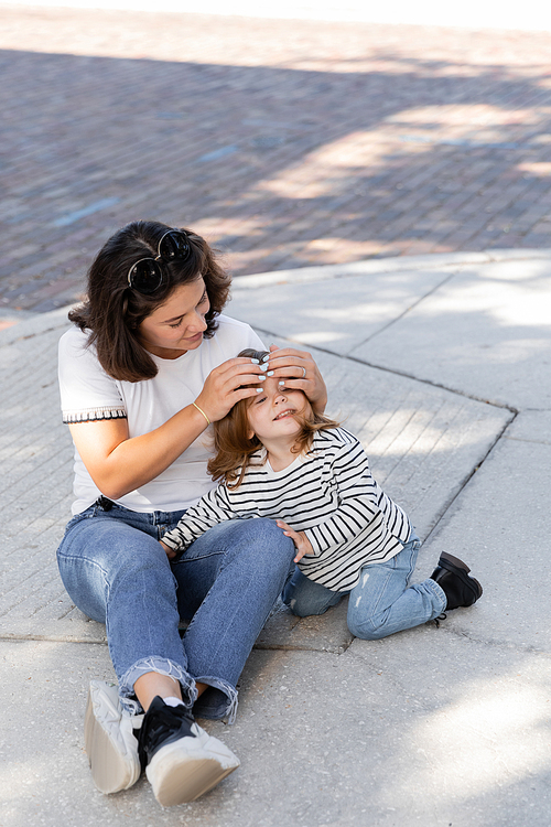 brunette mother in jeans sitting with excited daughter in casual clothes on street in Miami