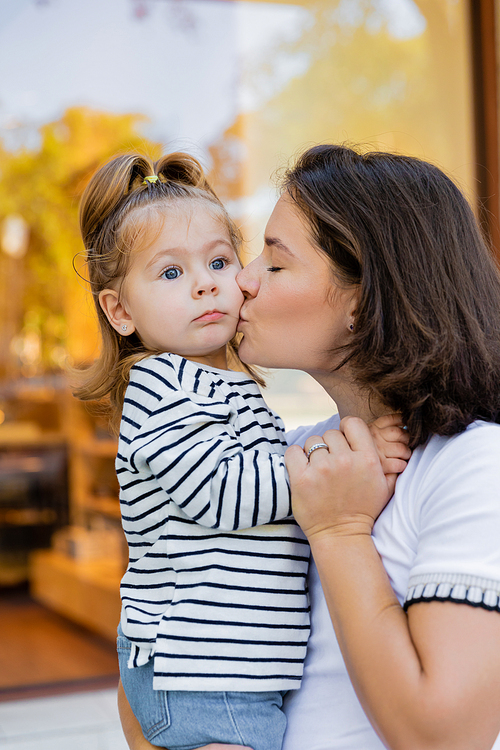 brunette mother kissing baby girl in striped long sleeve shirt near showcase of shop in Miami