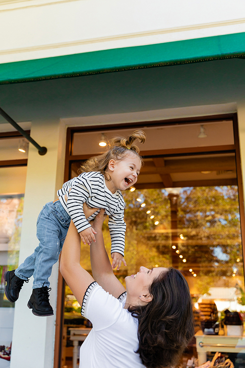 cheerful mother lifting happy baby girl near showcase of shop in Miami