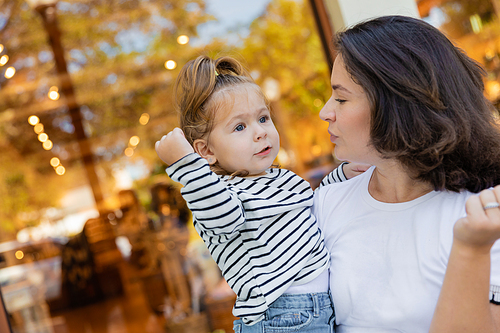 brunette mother holding in arms baby girl in striped long sleeve shirt near showcase of shop in Miami