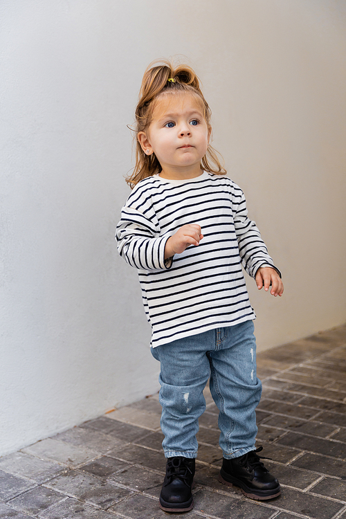 full length of toddler baby girl in striped long sleeve shirt and jeans standing near white wall