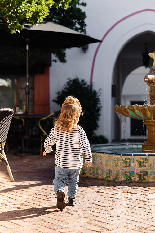 back view of toddler baby girl in striped long sleeve shirt and jeans walking near fountain in Miami