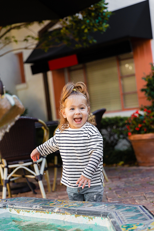 joyful toddler girl in striped long sleeve shirt standing near fountain in Miami