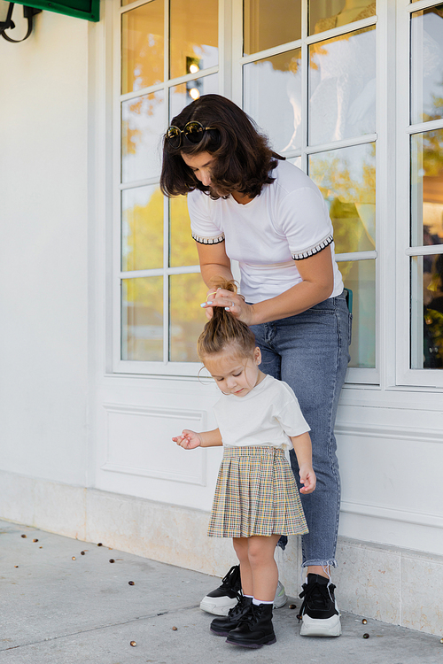 mother in jeans making hair of toddler daughter in skirt on porch of house