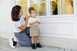 happy mother in jeans looking at toddler daughter in skirt near house