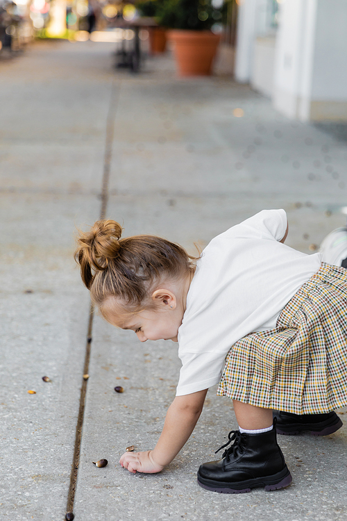 happy toddler girl in skirt picking up acorn from ground on street in Miami