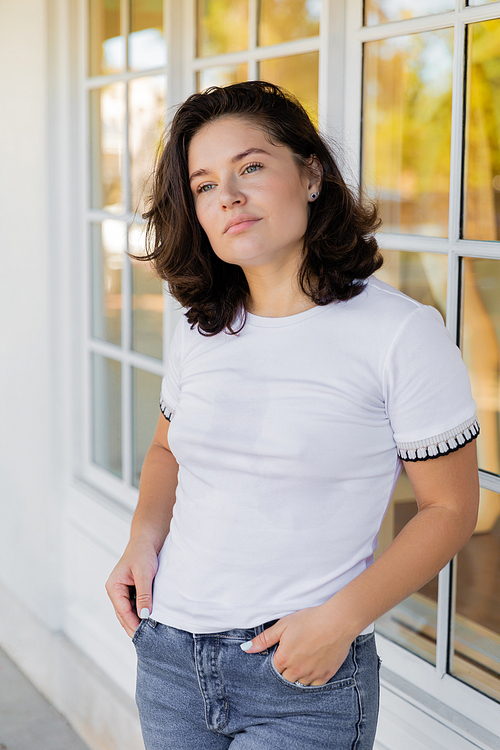 portrait of brunette woman in white t-shirt looking away while standing with hands in pockets near home