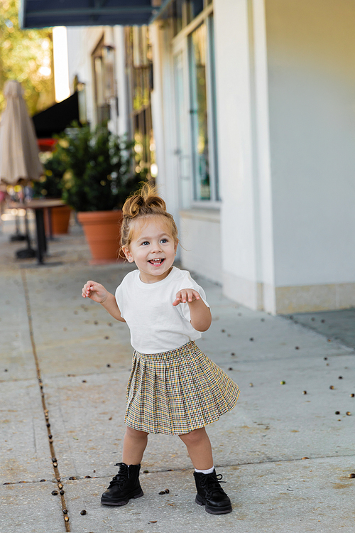 full length of positive toddler girl in skirt and white t-shirt standing on street in Miami