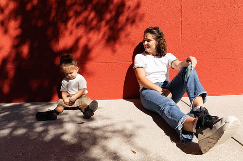 cheerful mother in jeans looking at toddler daughter while sitting near red wall