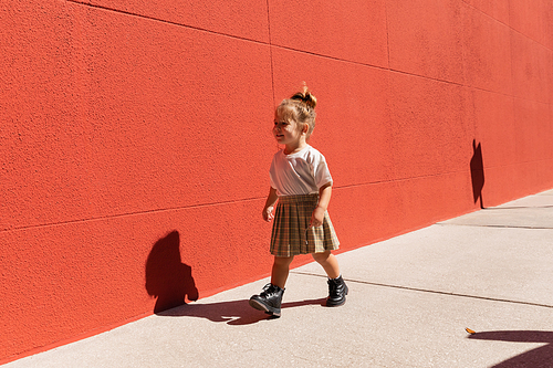 cheerful toddler girl in checkered skirt and white t-shirt walking near building with red wall