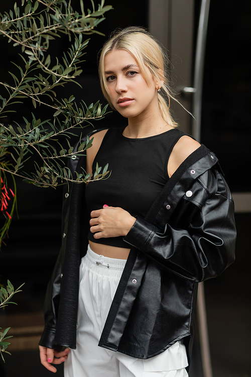 blonde woman in leather shirt jacket and black tank top standing near green plant