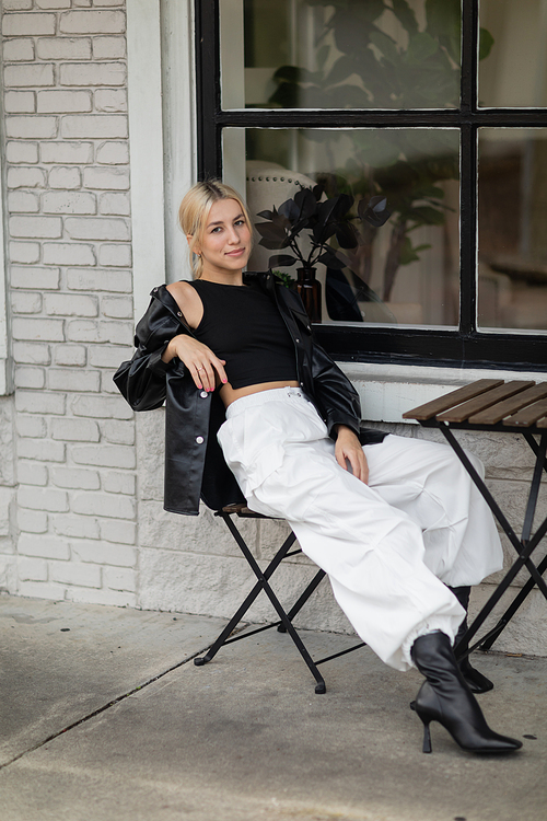 full length of blonde woman in leather shirt jacket and cargo pants sitting in outdoor cafe on street in Miami