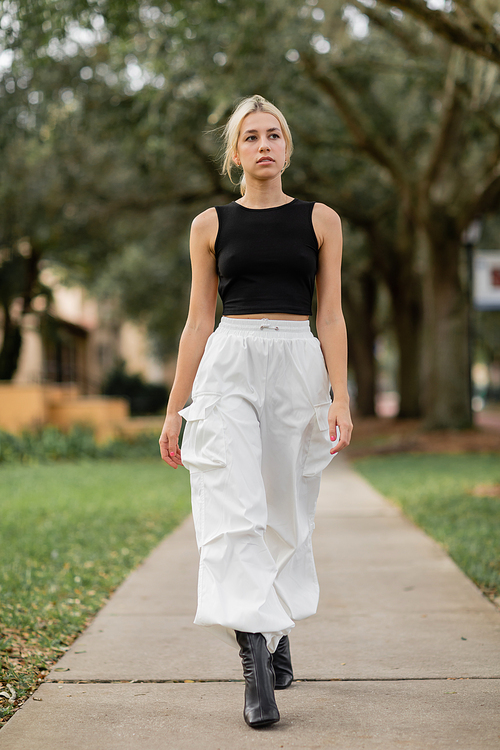 full length of young woman in white cargo pants and black tank top walking on green street in Miami